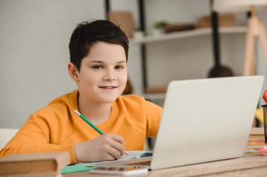 kalemle yazan gülümseyen çocuk ve evde okul işi yaparken kameraya bakmak