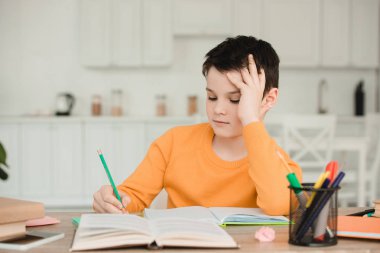 attentive schoolboy reading book and writing while doing schoolwork at home