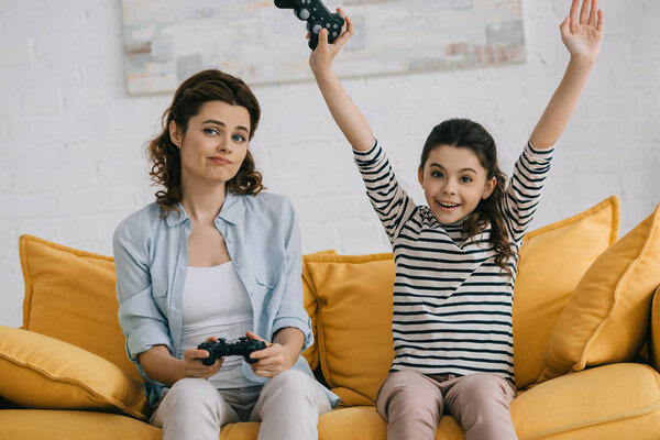 KYIV, UKRAINE - APRIL 8, 2019: Cheerful daughter showing yes gesture while sitting near upset mother holding joystick