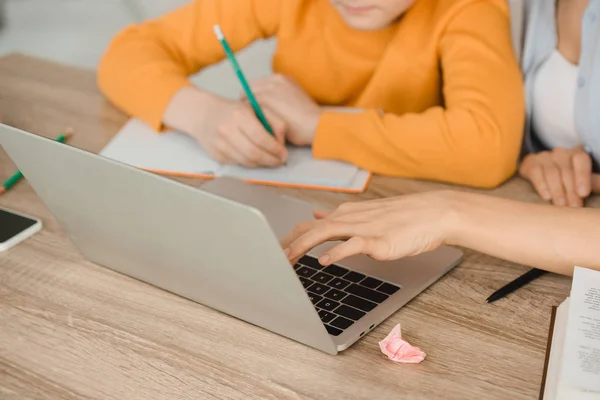 在家做功课时 使用笔记本电脑和男孩在抄本本上写作的妇女的裁剪视图 — 图库照片