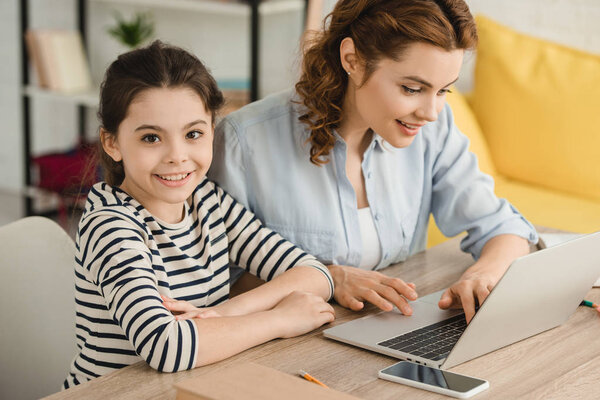 cheerful child looking at camera while using laptop together with mother