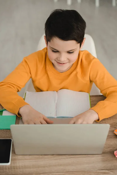 コピーブックを持って机に座り 自宅で学業をしている間 ノートパソコンを使って笑顔を見せた少年 — ストック写真