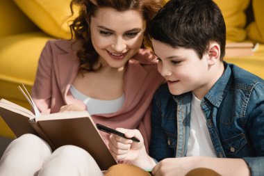 neşeli anne kitap tutan ve ev ödevi yapıyor sevimli oğlu yardım