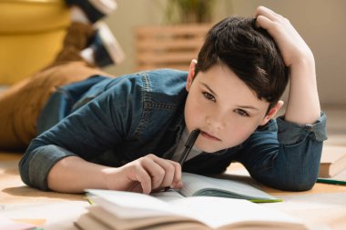 selective focus of thoughtful boy lying on floor near copy book while making schoolwork at home clipart
