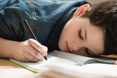 exhausted boy lying on floor and writing in copy book while making schoolwork at home clipart