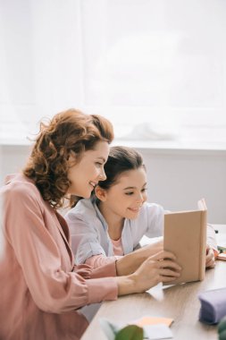 neşeli anne ve kızı masada oturan ve birlikte kitap okuma 
