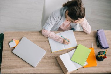 kitap, defter, dizüstü bilgisayar ve kalem kılıfı nın yanındaki ahşap masada otururken ev ödevi yapan okul çocuğunun genel görünümü