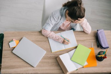 overhead view of schoolchild doing homework while sitting at wooden desk near book, notebooks, laptop and pencil case clipart