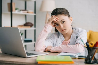 dizüstü bilgisayar ve dizüstü bilgisayarların yanında masada otururken kameraya bakan düşünceli çocuk