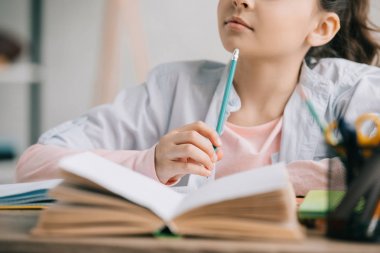 partial view of schoolkid holding pencil while sitting at desk near book and doing homework clipart