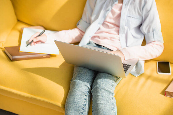 обрезанный вид ребенка, пишущего в копировальной книге и с помощью ноутбука, сидя на диване дома
