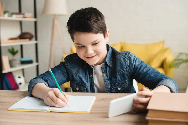 Ragazzo Sorridente Che Scrive Nel Quaderno Usa Smartphone Mentre Compiti — Foto Stock