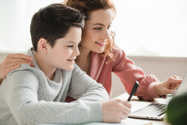 smiling woman hugging adorable son while doing schoolwork together at home