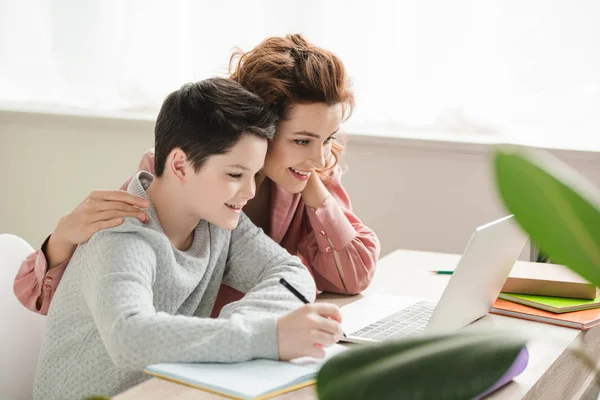 Feliz Madre Abrazando Adorable Hijo Mientras Haciendo Trabajo Escolar Juntos —  Fotos de Stock