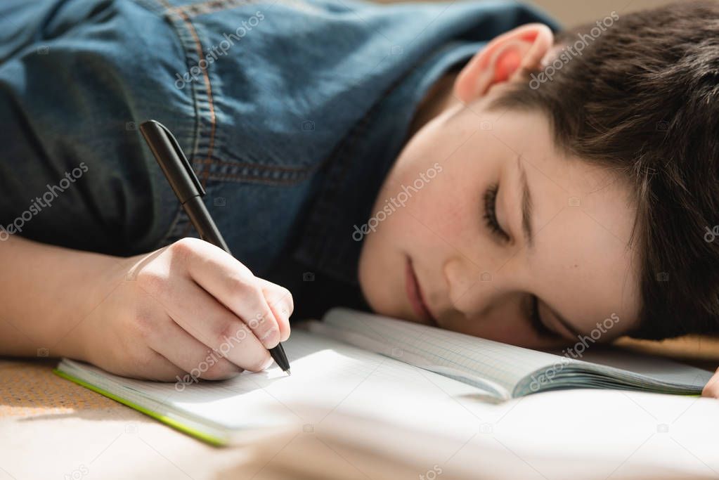 exhausted boy lying on floor and writing in copy book while making schoolwork at home