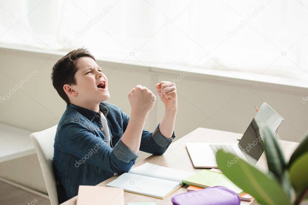 excited schoolboy showing yes gesture while sitting at desk and doing homework