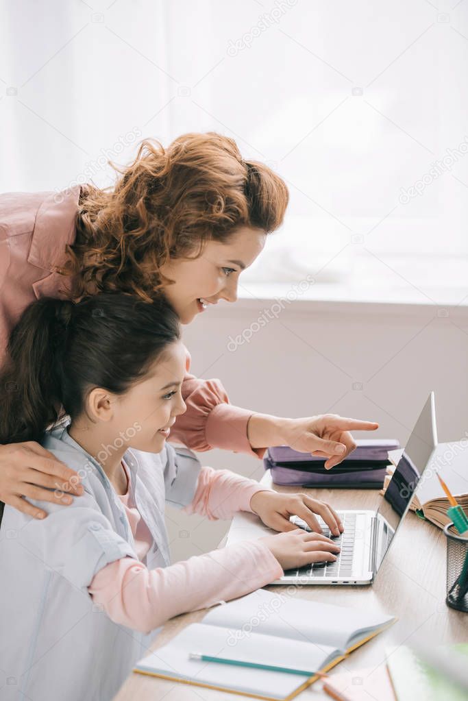 smiling mother pointing with finger at laptop screen near adorable daughter doing homework
