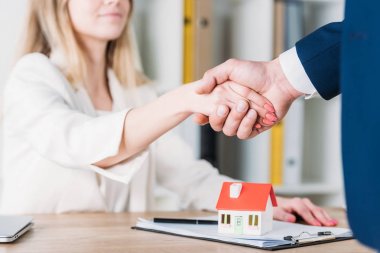 partial view of smiling woman shaking hands with realtor near house model on table clipart