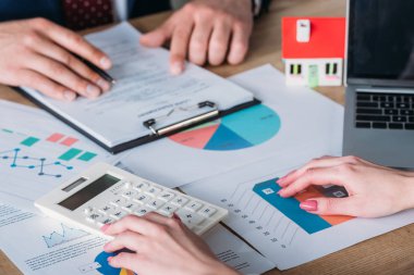 partial view of man studying loan agreement and businesswoman using calculator while sitting at workplace with house model, graphs and charts clipart