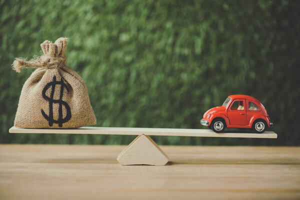 toy car and money bag with dollar sign balancing on seesaw on green background
