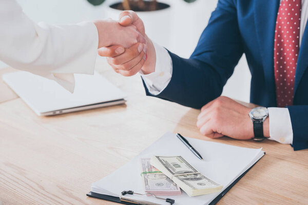 partial view of woman shaking hands with businessman near dollar banknotes on table