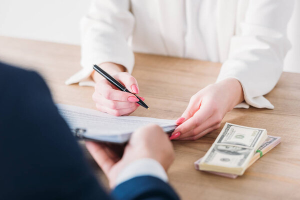 обрезанный снимок женщины, подписывающей контракт на планшете в руках менеджера возле банкнот доллара на столе
