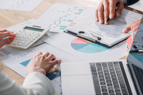 partial view of man studying loan agreement and businesswoman using calculator while sitting at workplace with graphs and charts