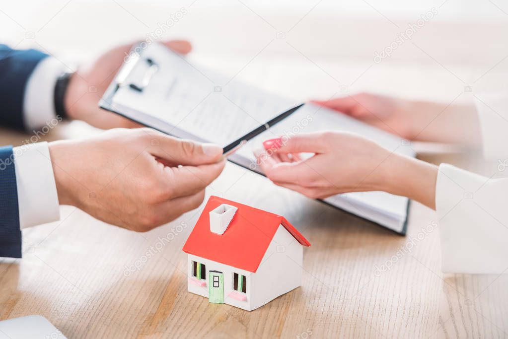 partial view of realtor giving pen and clipboard with loan agreement to client near house model 