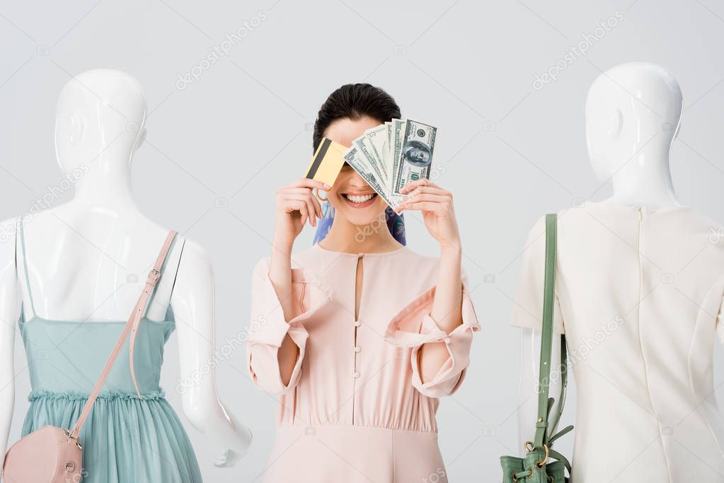 beautiful girl near mannequins covering face with money and credit card isolated on grey