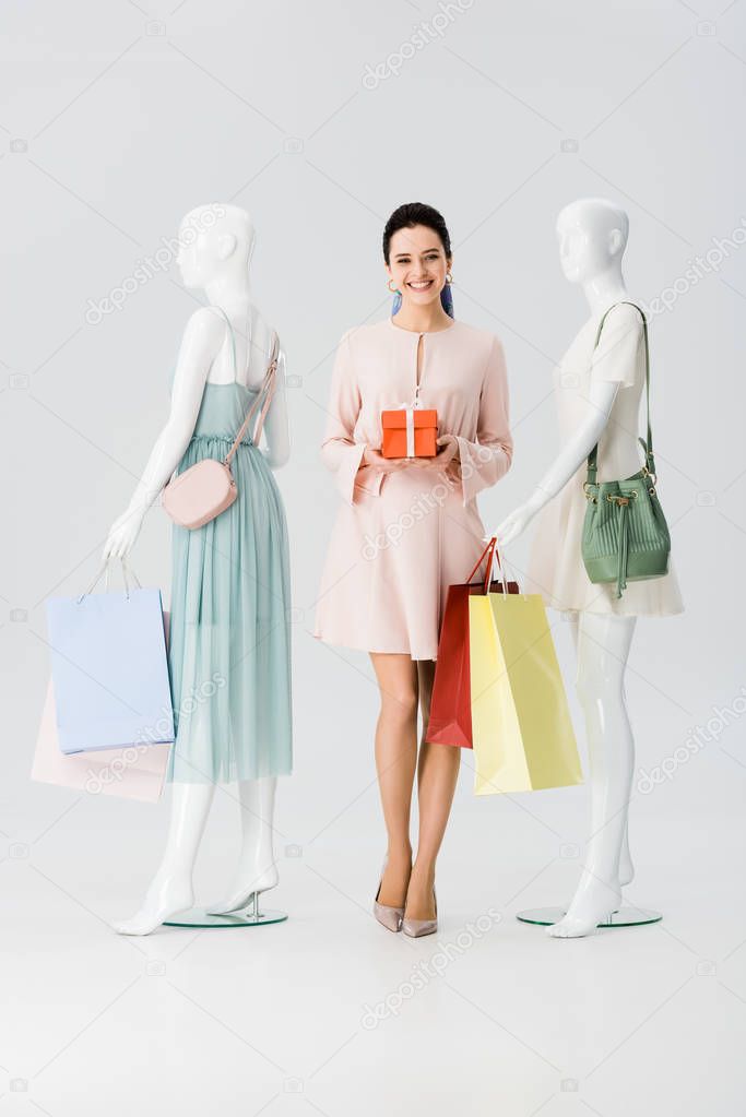 beautiful young woman with present near mannequins with shopping bags on grey