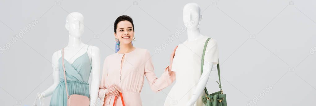 panoramic shot of beautiful girl posing with mannequins in dresses isolated on grey