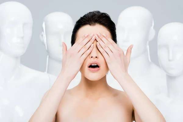 girl covering eyes while posing near mannequins isolated on grey