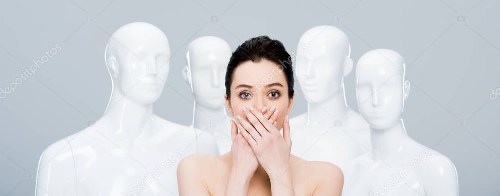 panoramic shot of girl covering mouth while posing near mannequins isolated on grey 