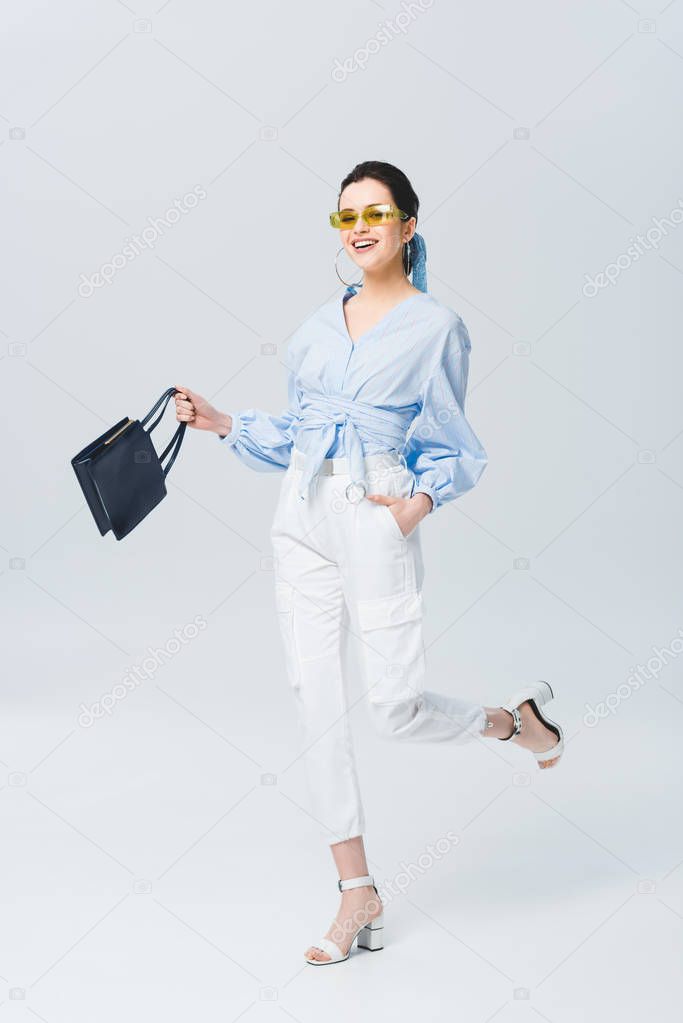 beautiful stylish young woman in sunglasses holding bag and smiling on grey