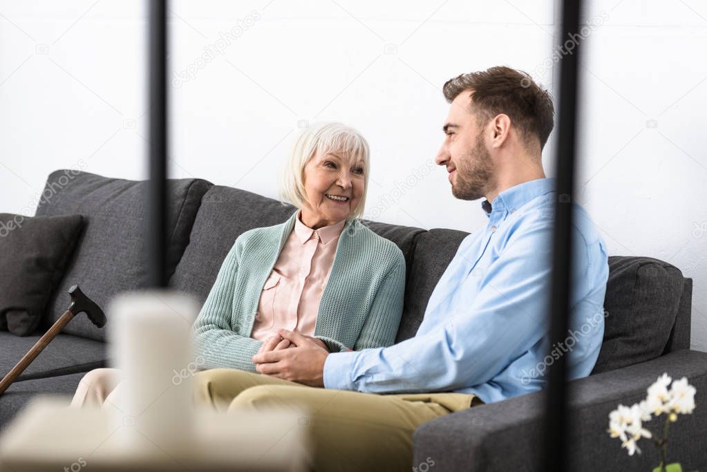 smiling senior mother and son holding hands and looking at each other while sitting on sofa