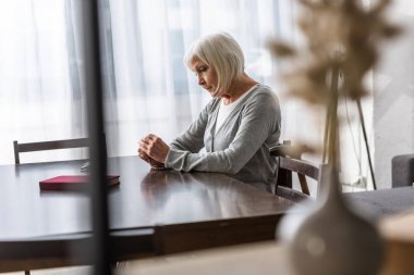 pensive senior woman sitting at table and holding rosary clipart