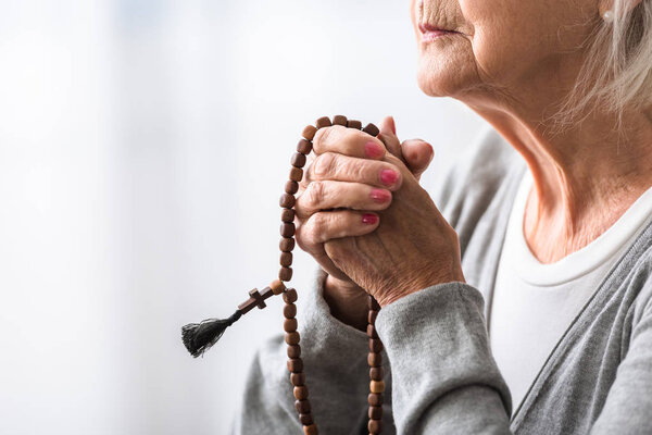 partial view of senior woman praying with wooden rosary 