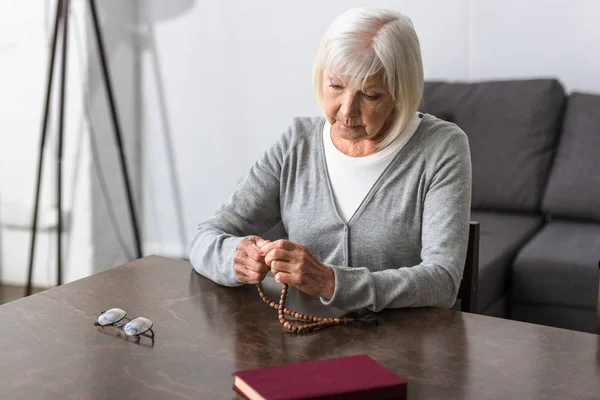 Pensivo Idosa Sentada Mesa Segurando Rosário — Fotografia de Stock