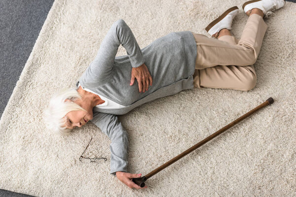 overhead view of sick senior woman with walking stick lying on carpet