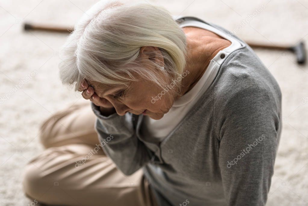 senior woman with migraine sitting on carpet and touching forehead with hand