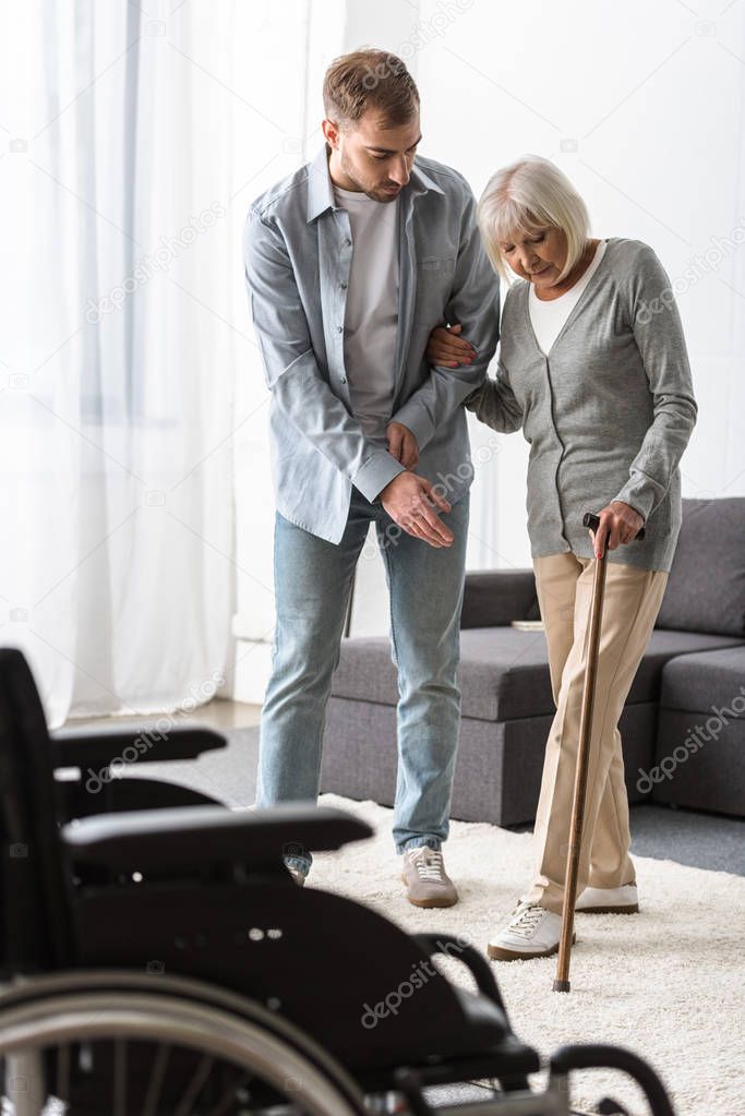 full length view of man helping senior mother with cane at home