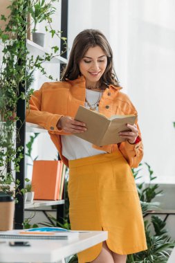 saksı bitkileri ile raf yakınında dururken neşeli kadın okuma kitabı