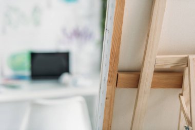 selective focus of wooden easel with fixed canvas and laptop with blank screen clipart