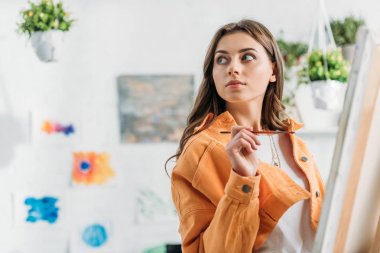 beautiful artist looking away while standing with paintbrush near easel clipart