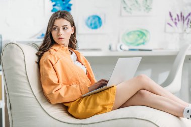 evde yumuşak şezlong üzerinde otururken dizüstü bilgisayar kullanan pensive genç kadın