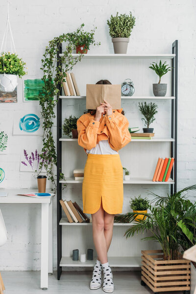 fashionable young woman standing bu rack and hiding face behind book