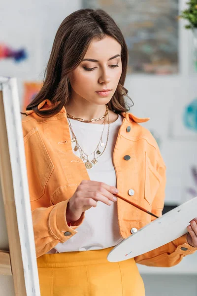 Attractive Pensive Artist Holding Palette Paintbrush While Standing Easel — Stock Photo, Image