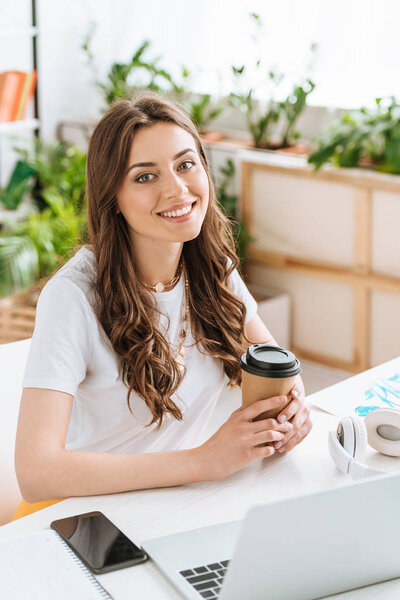 cheerful young woman looking at camera and holding paper cup while sitting at desk with laptop