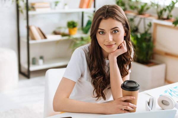 smiling young woman holding paper cup and looking at camera while sitting at desk at home