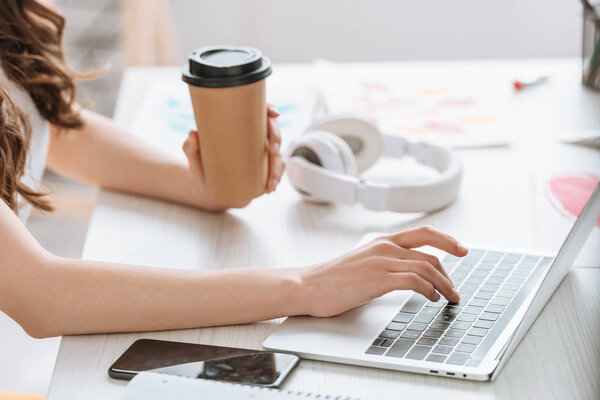 cropped view of young woman holding paper cup while using laptop near headphones